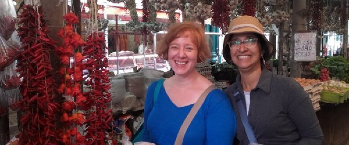 Two joyful women pose in front of a colorful market stall, enjoying their food tour at Porto's lively farm market.