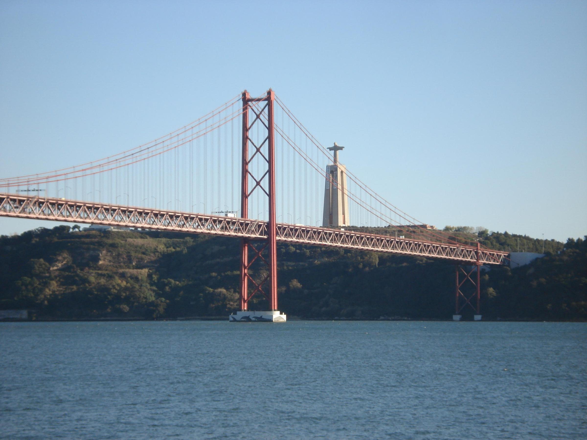 A view of the 25 de Abril Bridge spanning the Tagus River in Lisbon, with the city skyline and calm waters visible beneath.
