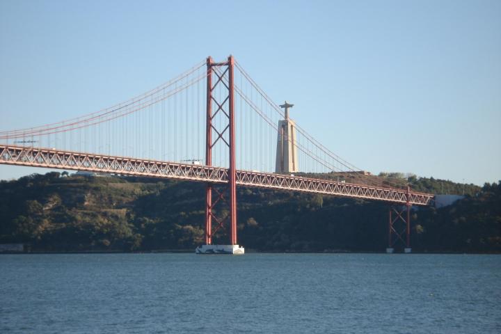 A view of the 25 de Abril Bridge spanning the Tagus River in Lisbon, with the city skyline and calm waters visible beneath.