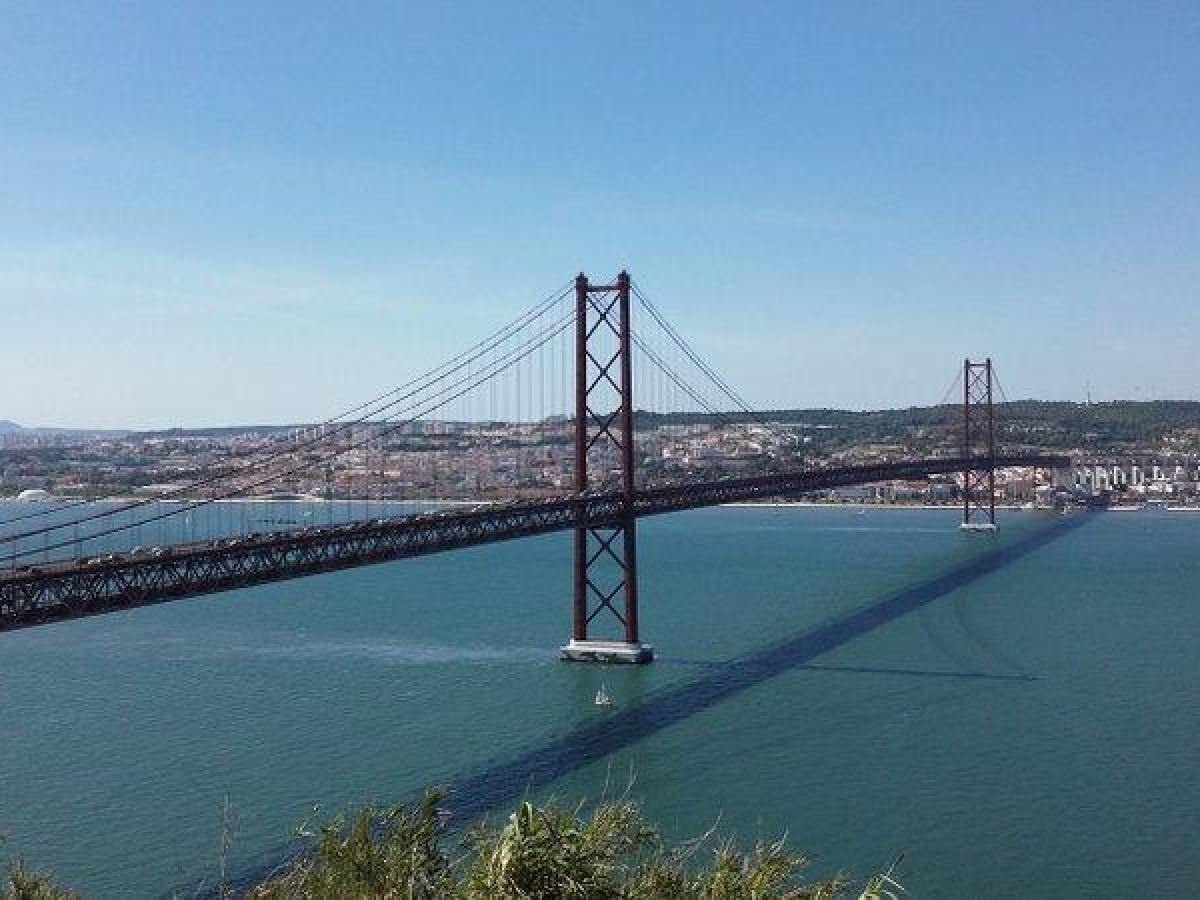 Lisbon's 25 de Abril Bridge gracefully spans the Tagus River, reflecting in the water beneath a bright, cloud-dotted sky.
