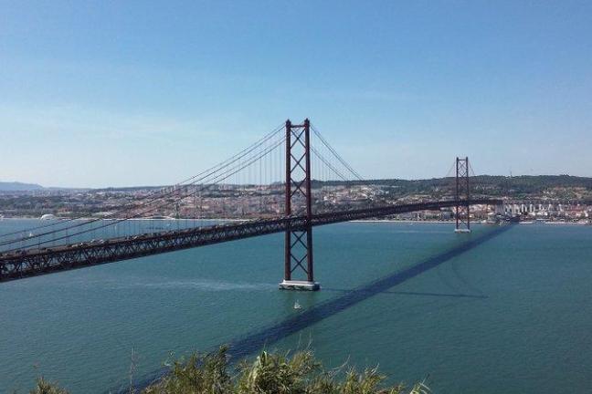 Lisbon's 25 de Abril Bridge gracefully spans the Tagus River, reflecting in the water beneath a bright, cloud-dotted sky.