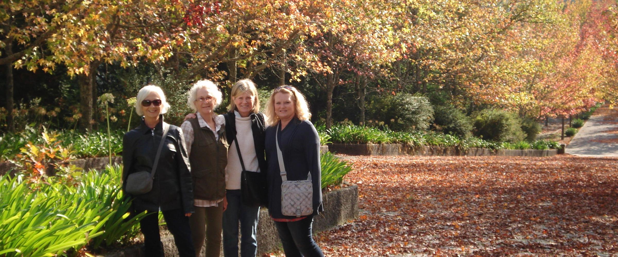Four women stand in a park, surrounded by fallen leaves, enjoying a hiking tour and the beauty of nature.