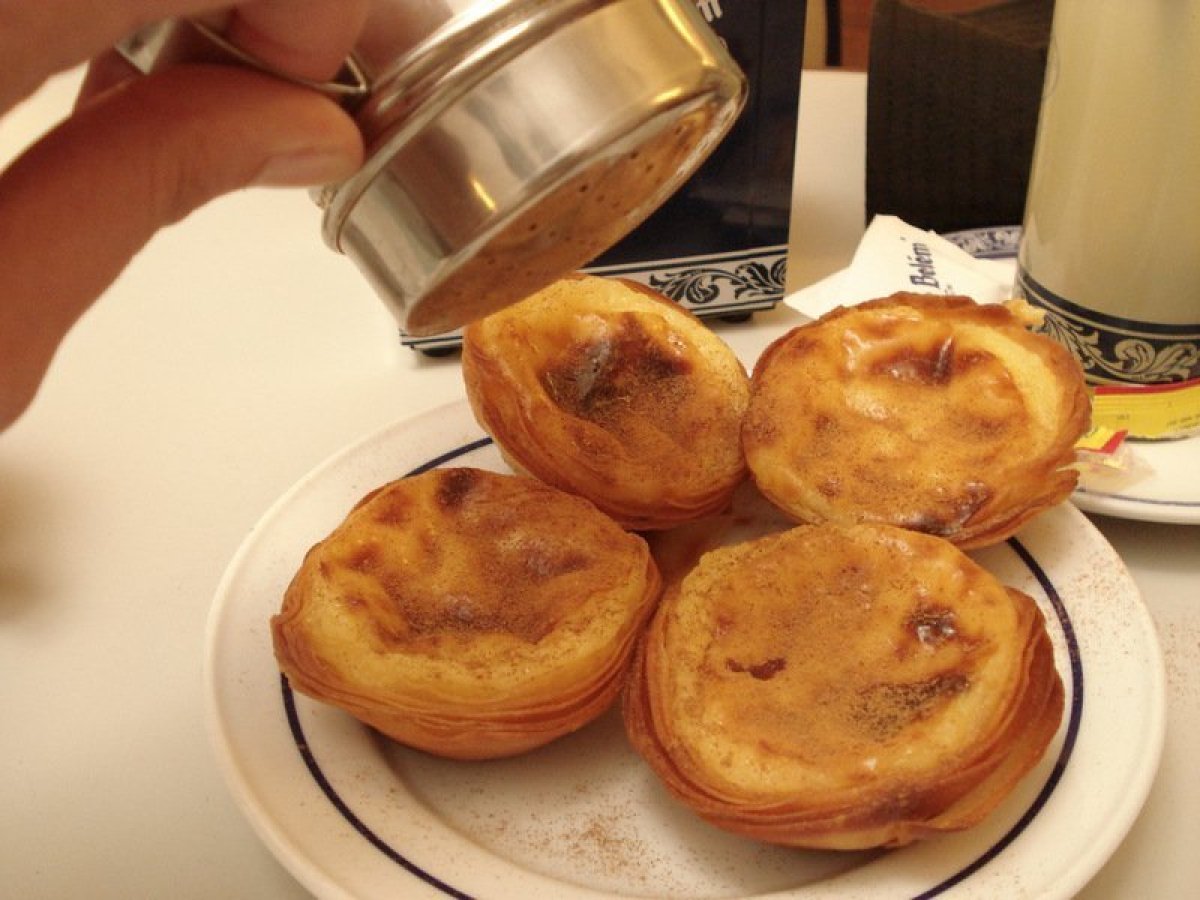 A hand delicately pours cinnamon into the tarts, crafting traditional Portuguese tarts with a perfect golden hue.