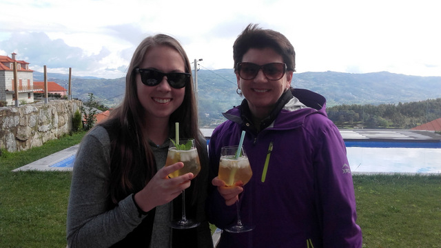 Two women toast with drinks, enjoying a stunning mountain view, one savoring a refreshing Portuguese sangria.