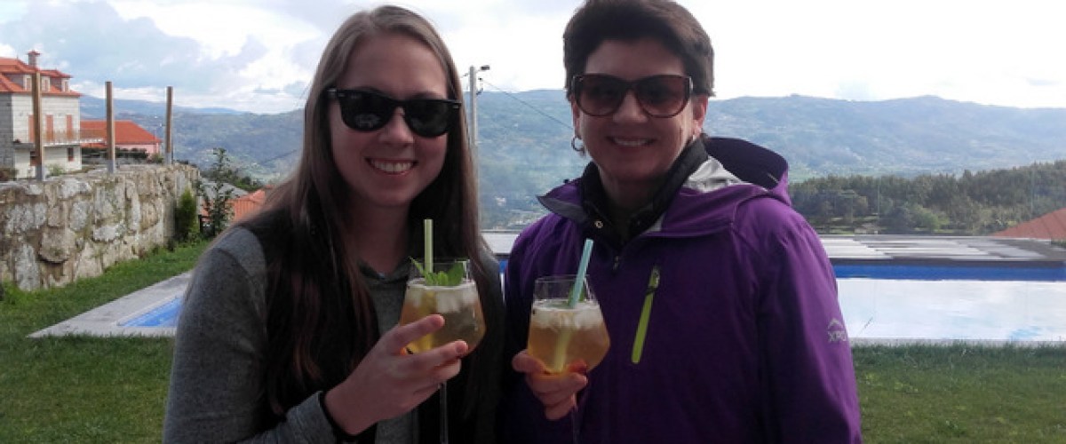 Two women toast with drinks, enjoying a stunning mountain view, one savoring a refreshing Portuguese sangria.
