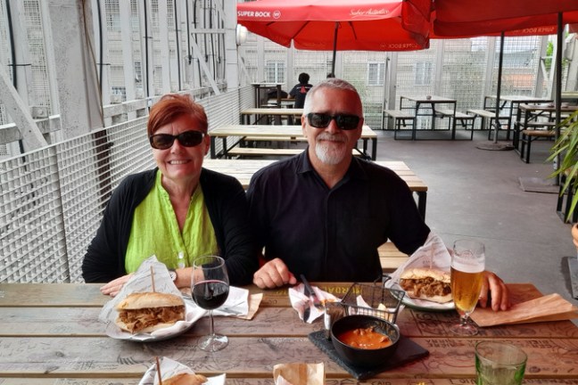 A couple enjoys a meal at a table, sharing delicious bifana sandwiches and gizzards together.