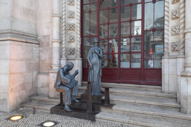 Statues of two individuals sitting on steps, embodying the spirit of a fado tour near a historic building.