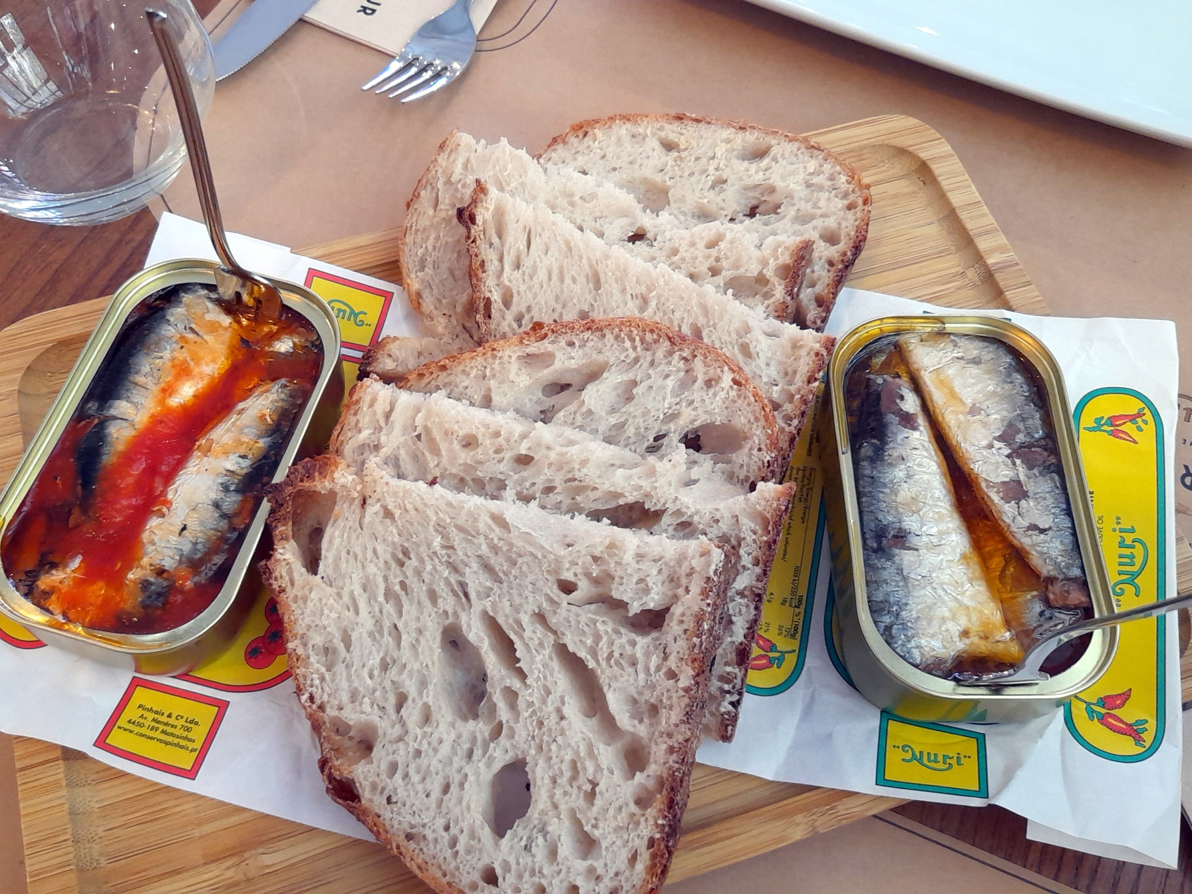 Two cans sit on a wooden cutting board, highlighting a delicious Portuguese food tour with sardines and bread.