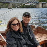 Two people seated on a boat on a river with a bridge and greenery in the background.