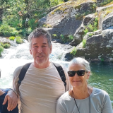 Two people smiling in front of a waterfall and rocky landscape.
