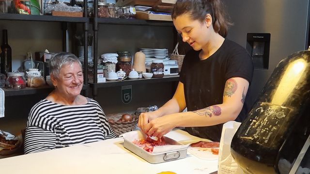 A woman prepares meat in a kitchen with an older woman, highlighting the rich flavors of Portugal's chourizo bread.