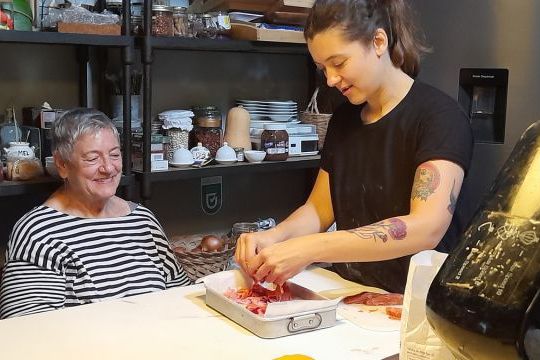 A woman prepares meat in a kitchen with an older woman, highlighting the rich flavors of Portugal's chourizo bread.