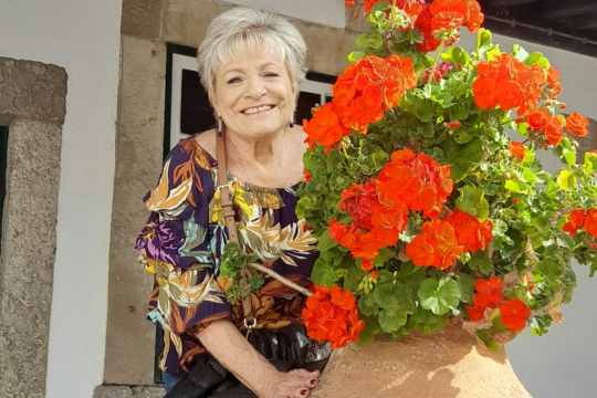 An older woman smiles as she holds a large pot of blooming flowers, enjoying springtime in Portugal during a small group tour.