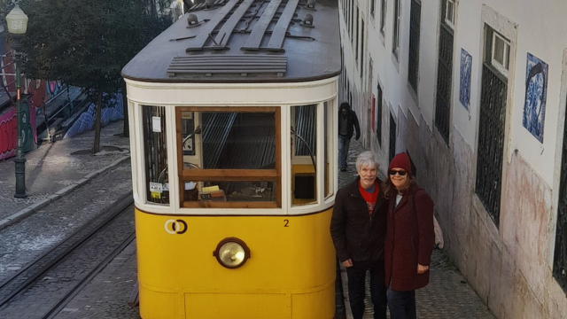 Two travelers stand next to a vibrant yellow tram in Lisbon, enjoying a walking tour with their friendly local guide.