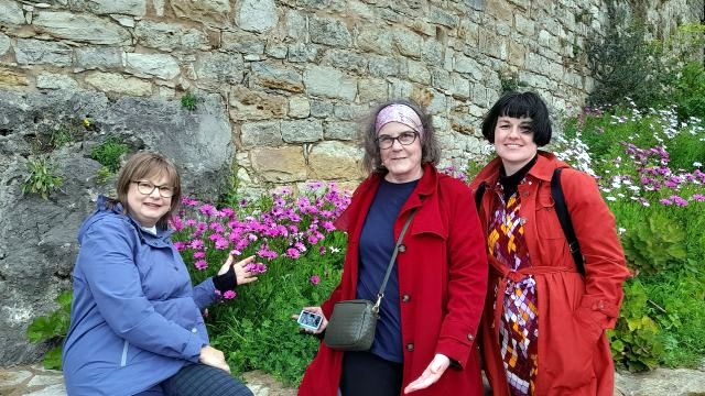 A small group of three women poses by a flowered stone wall while touring Obidos, Portugal.