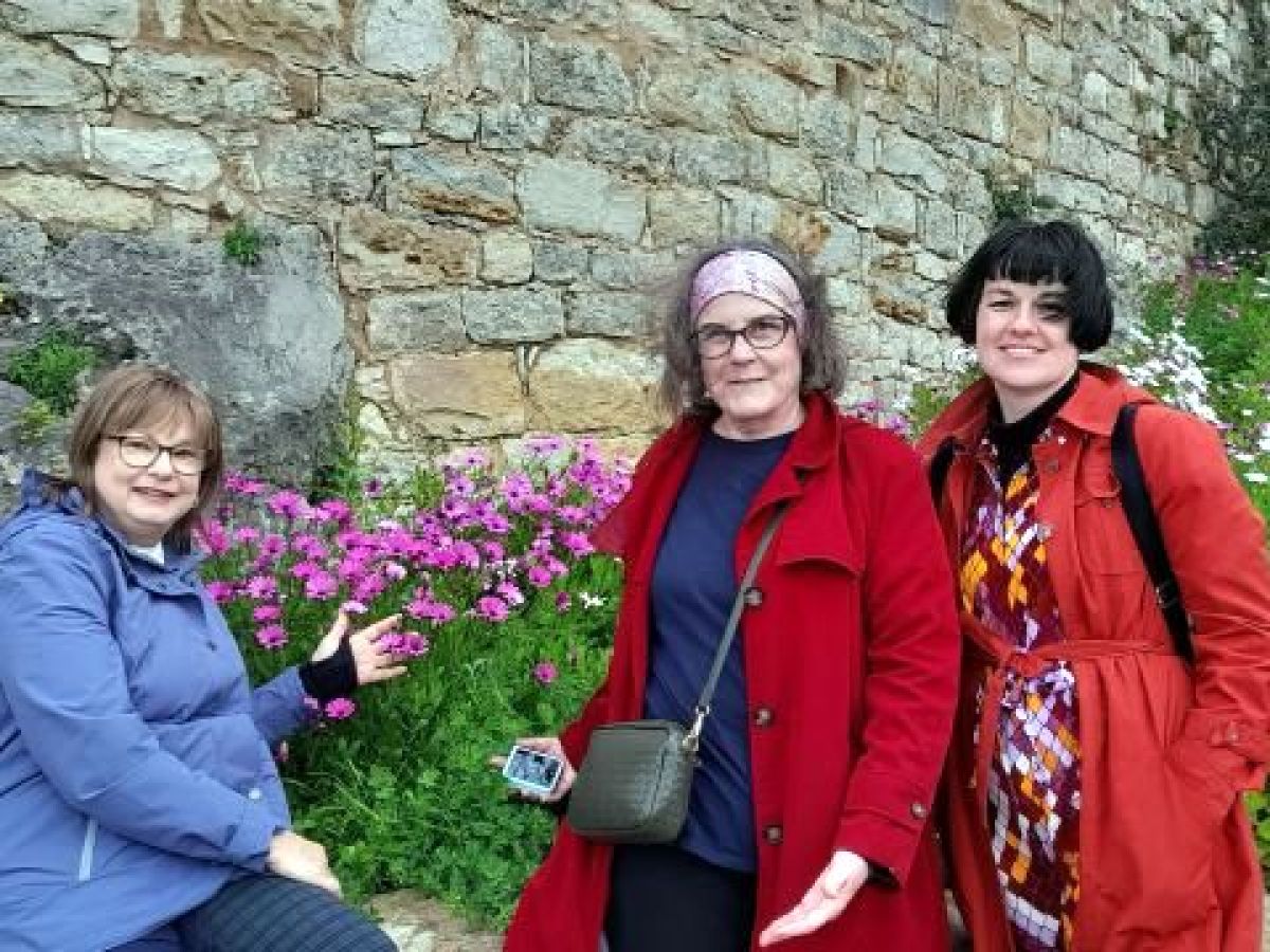 A small group of three women poses by a flowered stone wall while touring Obidos, Portugal.