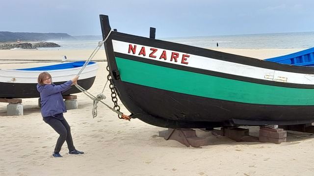 A woman stands beside a boat on Nazaré beach, capturing a humorous moment in a lively coastal setting.