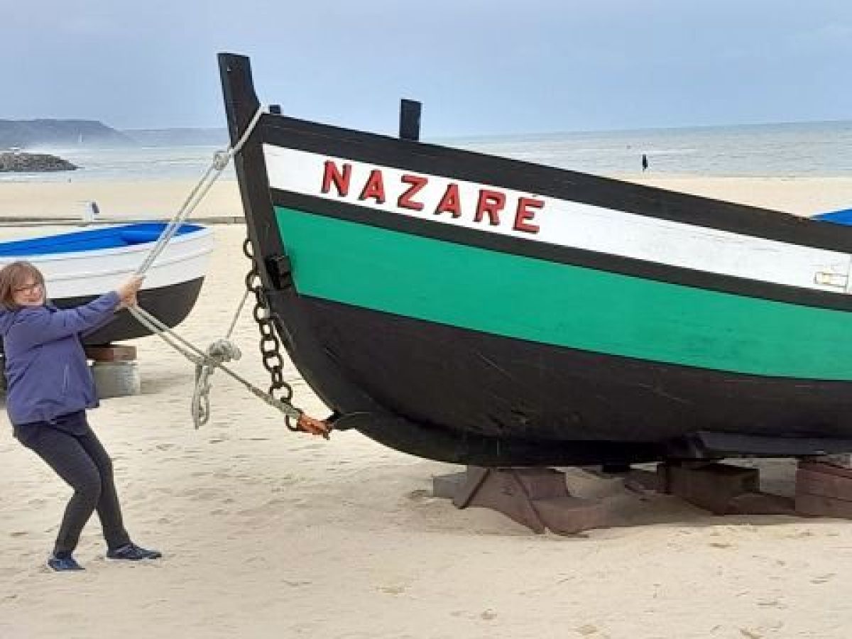A woman stands beside a boat on Nazaré beach, capturing a humorous moment in a lively coastal setting.