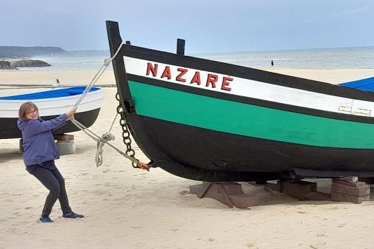 A woman stands beside a boat on Nazaré beach, capturing a humorous moment in a lively coastal setting.