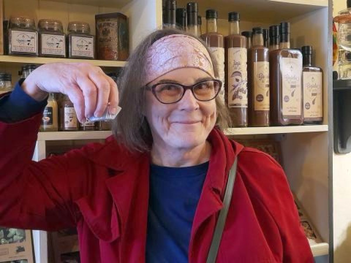 A woman wearing glasses and a red coat holds a bottle of Ginja liqueur, smiling at the camera.