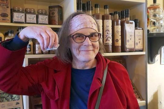 A woman wearing glasses and a red coat holds a bottle of Ginja liqueur, smiling at the camera.