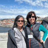 Two women in sunglasses standing on a bridge with a city and river in the background.