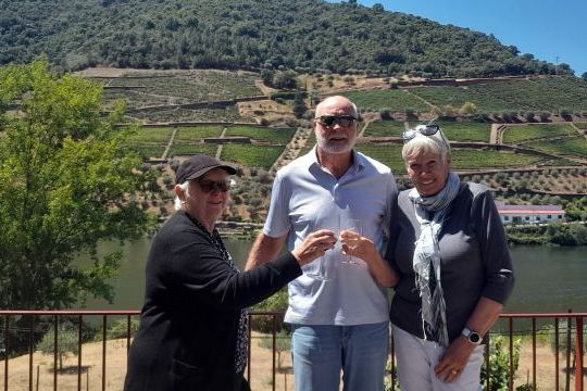 A small group enjoys wine by a river, with lush vineyards of the Douro Valley visible behind them.