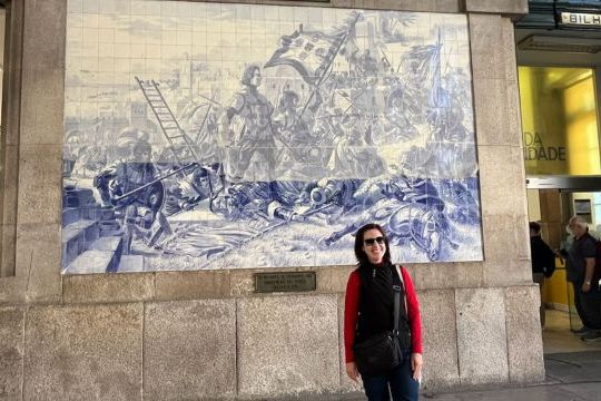 A tour guide woman poses in front of a large blue and white mural at São Bento train station in Porto.