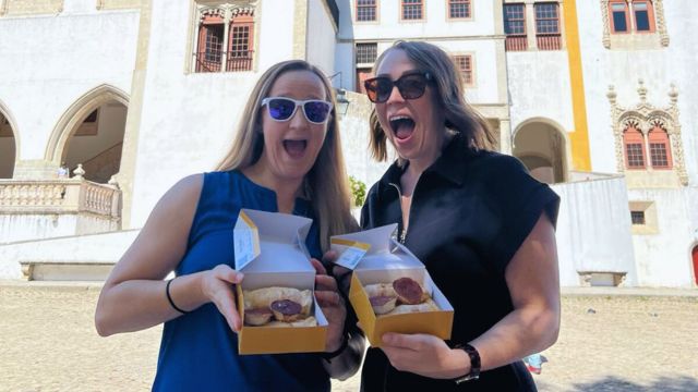 Two women display local cakes in front of a palace during a food tour in Sintra, Portugal.