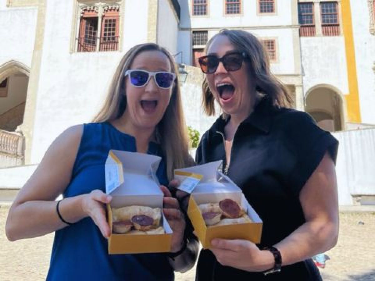 Two women display local cakes in front of a palace during a food tour in Sintra, Portugal.
