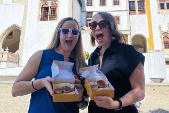 Two women display local cakes in front of a palace during a food tour in Sintra, Portugal.